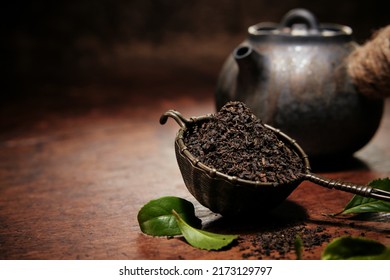 Heap of dried loose tea in metal ladle placed on wooden table with tea teapot in kitchen during teatime preparation