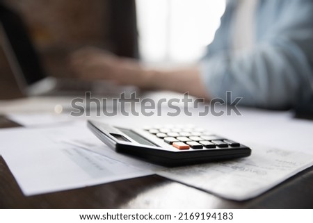Heap of papers, bills and calculator on desk close up, on background man working, pay bills using laptop. Accountant workflow, finances management, taxes, business, e-bank application usage concept
