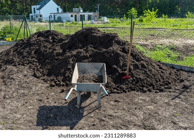 A heap of pure black earth lying in the yard next to the fence, visible shovel and empty wheelbarrow. - Shutterstock ID 2311289883