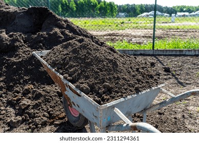 A heap of pure black earth lying in the yard next to the fence, visible shovel and full wheelbarrow. - Shutterstock ID 2311294361