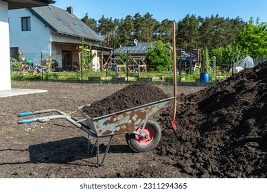 A heap of pure black earth lying in the yard next to the fence, visible shovel and full wheelbarrow. - Shutterstock ID 2311294365