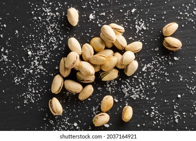 Heap of salted, roasted green pistachio nuts snack on black background with sea salt, healthy food snack, selective focus, flat lay top view from above