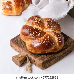 Heap of sweet round sabbath challah bread with white and black sesame seeds on small cutting board over white table with plastered wall at background. Square image with selective focus