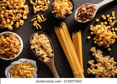 Heap Of Various Raw Pasta In Different Types And Shapes On Dark Brown Background. Overhead View. 