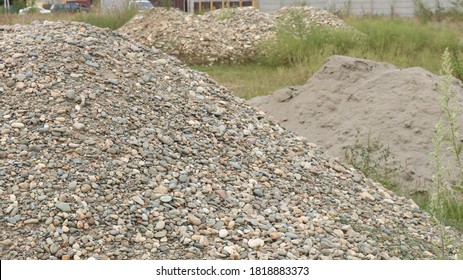 Heaps Of Large And Small Stones And Construction Sand Among A Grassy Landscape With Several Cars In The Background Of The Street, Embankments Of Building Bulk Components On The Site For Construction 