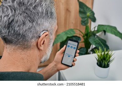 Hearing impaired mature man adjusts settings for his BTE hearing aid via smartphone. Hearing aids, deafness treatment, innovative technologies at audiology - Shutterstock ID 2105518481