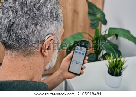Hearing impaired mature man adjusts settings for his BTE hearing aid via smartphone. Hearing aids, deafness treatment, innovative technologies at audiology