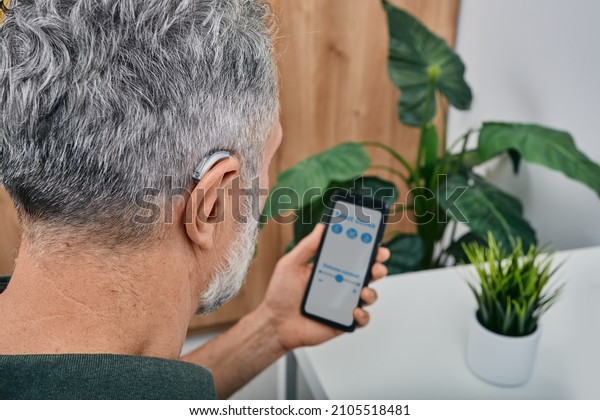 Hearing impaired mature man\
adjusts settings for his BTE hearing aid via smartphone. Hearing\
aids, deafness treatment, innovative technologies at\
audiology