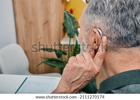 Hearing solutions for elderly deafness people. Older gray-haired man tunes his hearing aid behind the ear by pressing his finger on setting button while visit to hearing clinic