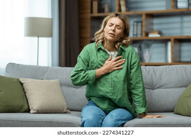 Heart attack, panic attack,. An elderly woman sits on the sofa at home and holds her hand to her chest. It is difficult to breathe, feels severe pain.