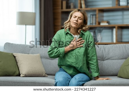 Heart attack, panic attack,. An elderly woman sits on the sofa at home and holds her hand to her chest. It is difficult to breathe, feels severe pain.