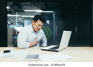 Heart attack at work. A young Asian man, businessman, office worker holds his heart, feels pain, had a stroke, heart attack at the desk in the office.