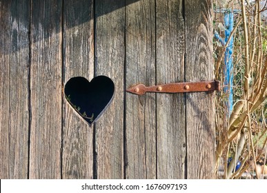 Heart Cutout Window In The Wooden Planks Of A Vintage Outdoors Toilet With A Rusty Door Hinge Pointing At It And An Ivy Plant Lurking For Sunlight.