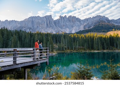 In the heart of the Dolomites, two hikers stand on a wooden deck, captivated by the turquoise lake and impressive mountain peaks. Lake Carezza or Karersee Dolomites in Italy. - Powered by Shutterstock