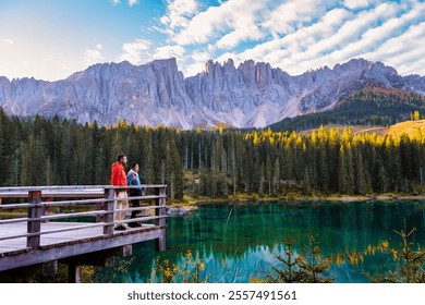 In the heart of the Dolomites, two hikers stand on a wooden deck, captivated by the turquoise lake and impressive mountain peaks. Lake Carezza or Karersee Dolomites in Italy at Autumn - Powered by Shutterstock