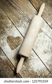 Heart drawn in sprinkled cooking flour. Wooden rolling pin with remnants of flour in a rustic kitchen on an old grainy textured wood surface