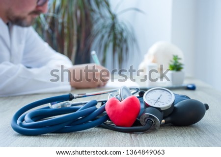 Heart health and diagnosis and treatment of cardiac disease concept photo. In foreground figure of red heart surrounded by stethoscope on table in background - blurry figure of doctor prescribing drug