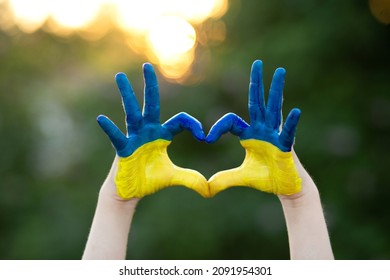 Heart Shape With Child Hands Of Kid Body Language For Children Love, Kindness, Patriot Love Concept. Heart Hand Over Sunset Sky Bokeh Background Painted In Ukrainian Flag Colors