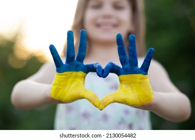 Heart Shape With Child Hands Of Kid Body Language For Children Love, Kindness, Patriot Love Concept. Heart Hand Over Sunset Sky Bokeh Background Painted In Ukrainian Flag Colors