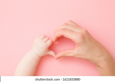 Heart shape created from infant and young mother hands on light pink table background. Pastel color. Lovely emotional, sentimental moment. Love, happiness and safety concept. Closeup.