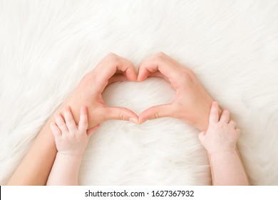 Heart shape created from young woman fingers. Infant hands on mother arms. White, soft, fluffy fur carpet background. Lovely emotional, sentimental moment. Closeup. Point of view shot.
