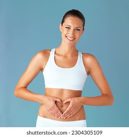 Heart shape, hand gesture on stomach and woman in portrait, gut health and wellness isolated on blue background. Weight loss, fitness and healthy female person with self love and body care in studio