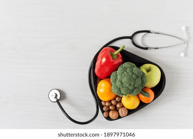Heart shaped bowl with vegetables, fruits, nuts and stethoscope on white wooden background top view. Healthy eating concept.