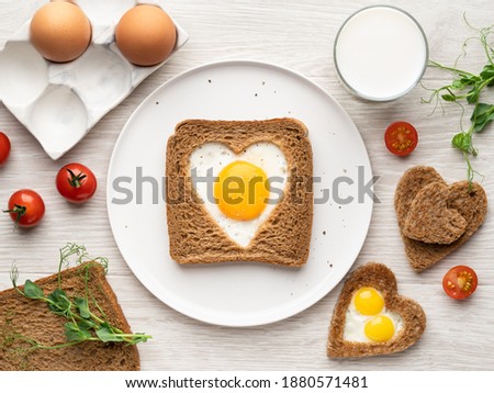 Heart shaped egg in tosted bread on white ceramic plate. Valentine's day concept. Love breakfast design. Homemade healthy sandwich, glass of milk, eggs and microgreens. Festive morning table. Top view