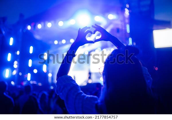 Heart shaped hands at concert, loving the artist\
and the festival. Music concert with lights and silhouette of\
people enjoying the\
concert.