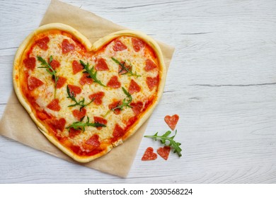 Heart shaped Italian pizza with pepperoni,arugula,pizza sauce and mozzella cheeses on parchment paper with white wood table background.Love concept for Valentine's day.Top view.Copy space

