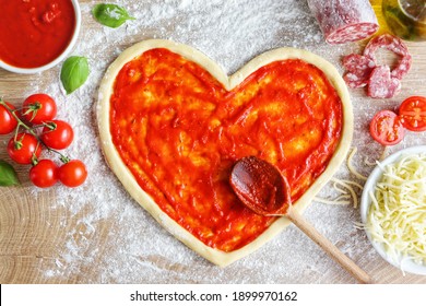 Heart Shaped Pizza Dough Covered With Pizza Sauce On Wooden Table  With Cherry Tomatoes,tomato Sauce,mozzarella Cheeses,salami,olive Oil And Basil Leaves.Cooking With Love Concept For Valentine's Day