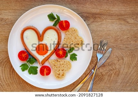 Heart shaped sausage with fried egg served with heart shaped bread and tomatoes on plate with white wood background.Romantic art food idea for Valentine's breakfast.Top view.Copy space

