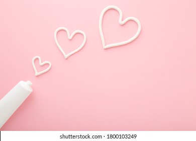 Heart Shapes Created From Cream. White Tube On Light Pink Table Background. Pastel Color. Care About Face, Hands, Legs And Body Skin. Closeup. Empty Place For Text Or Logo.