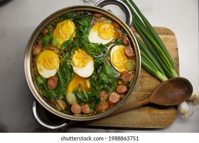 A Heart Warming Comfort Food - Noodle Soup With Well-cooked Boiled Eggs, Top With Chicken Sausages And Spinach, In A Pot With A Wooden Ladle And Fresh Spring Onions At The Side