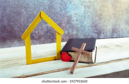 Heart and Wooden cross leaning on a bible, next to small yellow house wooden on white background. copy space