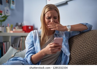 Heartbroken blond girl holding pink smartphone while sitting on couch. She looking at phone display with unhappy expression and covering mouth with hand as if going to cry