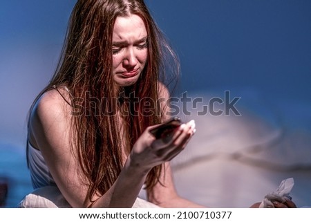 Heartbroken. Get the bad news. Get upset. Depression. Broken heart. A woman with a face dirty from mascara sits in bed and cries, holding a phone in her hands. To suffer.