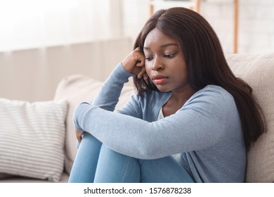 Heartbroken. Upset african american girl sitting on couch at home, with thoughtful face expression, feeling lonely and misery