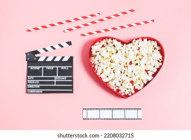 A heart-shaped box with popcorn, a clapperboard, straws and a film strip lie on a pink background, top view. Cinema and valentine day concept.