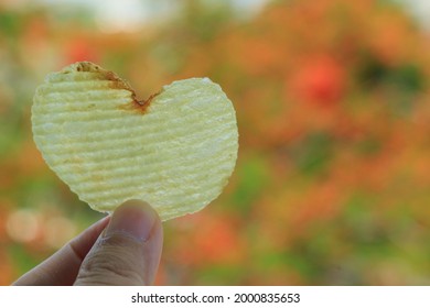 Heart-shaped potato chip in woman hand with colorful leaves in autumn background. Food and Valentine's day concept.