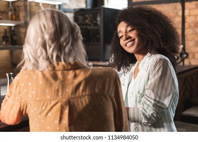 Heart-warming conversation. Beautiful curly woman sitting next to her best friend at the bar counter and chatting with her while smiling fondly