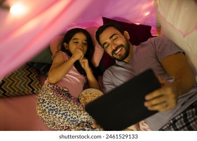 Heartwarming image of a father and daughter under a blanket fort at home, enjoying movie night on a tablet  - Powered by Shutterstock