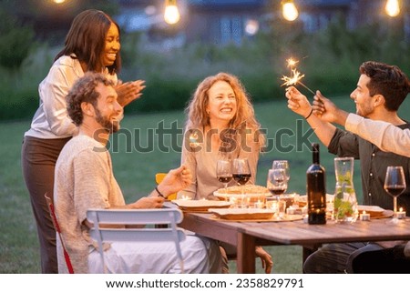 A heartwarming scene unfolds as a diverse group of millennial friends gathers to celebrate the birthday of a lovely Caucasian woman. In the enchanting evening ambiance illuminated by strings of lights