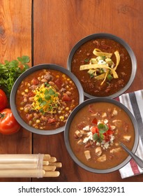 Hearty Soup Trio With Mexican Tortilla, Texas Chili And Chicken Enchilada Soup In Grey Bowls On A Warm Wood Background Served With Bread Sticks In A Vertical Overhead View