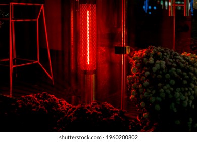 heating infrared stand for creating a comfortable temperature on the terrace of outdoor cafes and restaurants, a device glowing with red light near a glass window and flowers at night.