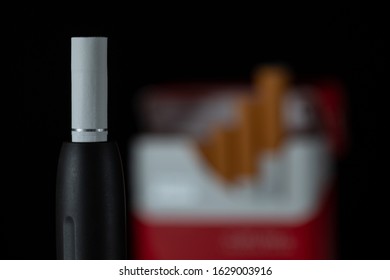 Heating Tobacco System On Black Background. Newest Electronic Cigarette. Heat Not Burn Tobacco Products. Iqos Heating Tobacco System In Front Of Blurred Cigarette On Black Background