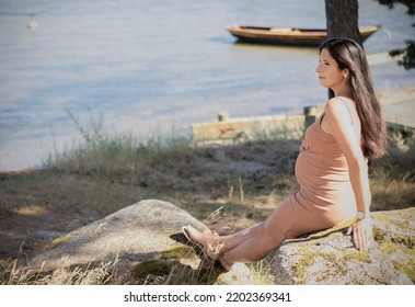 Heavily Pregnant Attractive Brunette Woman Sitting On A Big Rock By The Seaside With A Fishing Boat Behind Her On The Sea On A Sunny Summer Day