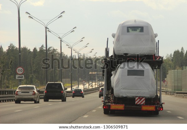 Heavy car carrier truck transports new cars in\
protective covers on semi-trailer on suburban asphalt highway road\
on summer day against forest and blue sky - logistics business in\
Europe, delivery
