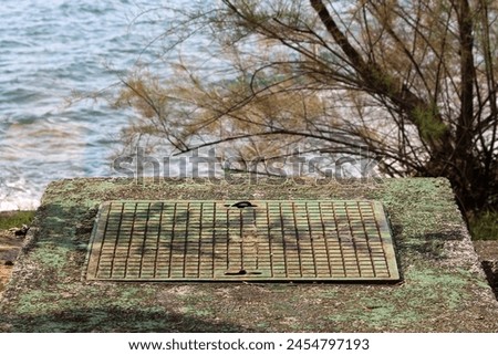 Heavy closed rectangular metal manhole cover with concrete foundation covered with thin layer of green moss with trees and sea in background on warm sunny autumn day