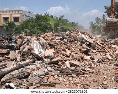 A heavy construction machine clears a large heap of broken bricks, concrete debris, and twisted metal at an urban demolition site under a bright sunny sky.